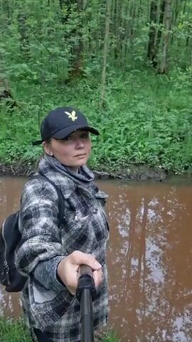 A woman stands at the edge of a muddy river or stream in a forest, holding a selfie stick. She has fair skin, and wears a black baseball cap with a yellow logo, a gray and black plaid jacket, and a gray scarf. She also wears a black backpack. The forest is lush with green vegetation and tall trees. The water in the river is murky brown and reflects the surrounding trees. The woman is looking directly at the camera.
