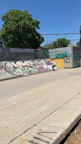 A daytime video shows a person riding a scooter in a concrete skatepark under a bright, clear sky. The rider, wearing a white t-shirt, gray pants, and an orange helmet, is performing a trick on a quarter pipe. They ride up the ramp, go over the coping, and then come back down, landing smoothly.

The skatepark features concrete ramps and walls, many of which are covered in colorful graffiti. Visible graffiti includes words like "PAT," "FOREVER," "SHO," "NIKO LUXITO FELBR," "MILO," and "ERIK," along with various cartoon-like characters and abstract designs. In the background, green trees are visible, and distant hills can be seen beneath the blue sky. The overall scene suggests a sunny day at an urban skatepark in Esplugues de Llobregat, Spain.