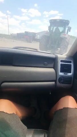 The video shows a view from inside a vehicle, looking out through the windshield at a tractor. The tractor is green and has large tires, and it is moving away from the camera. In the background, there are several buildings and a cloudy sky. The time of day appears to be daytime, and the weather seems to be sunny with some clouds. The scene looks like it could be a rural or industrial area. There are no visible people in the foreground or clearly identifiable text in the video. The overall impression is of a utilitarian, possibly agricultural or construction setting.