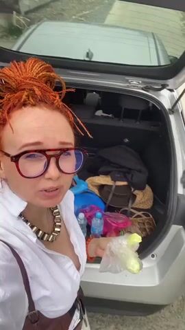 A group of three people are gathered around a car with its trunk open. The person in the foreground, a woman with bright orange dreadlocks, glasses, and a white shirt, is holding a clear plastic bag containing cucumber slices and is about to eat one. To her right, a person wearing a large straw hat is looking at their phone. Behind them, another person in a light-colored romper and white cap is holding and eating an apple. The car appears to be parked on the side of a road with trees and buildings in the background. The overall setting suggests a casual stop during a trip.