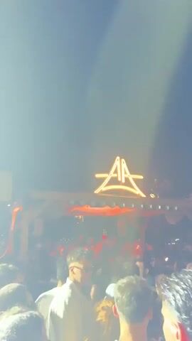 This is a vertical video showing a crowd of people at an outdoor nighttime event. A large, illuminated logo resembling a star with the letters "AA" inside is visible above a raised structure with lights. Many people are gathered below, some are dancing, and the atmosphere appears to be lively. The lighting is dynamic, with bright flashes and a general nighttime ambiance.