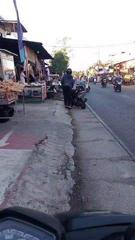 A person wearing a dark jacket and helmet is standing next to a parked motorcycle on a sidewalk. In the background, there are several stalls selling goods, possibly food items, and more motorcycles and people are on the road. The scene appears to be a street market with moderate traffic. The camera is mounted on a motorcycle, showing the handlebars and speedometer in the foreground. The time stamp is 00:00 to 00:04.