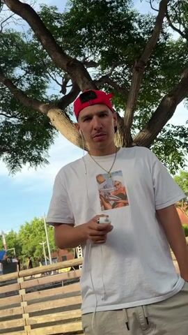 A male with a red baseball cap, a white t-shirt, a gold chain with a cross pendant, and white wired earbuds is standing outside under a tree. He is exhaling smoke from a white vape device held in his right hand. The t-shirt has a graphic of a woman in a bikini and a towel on her head, reading a newspaper.