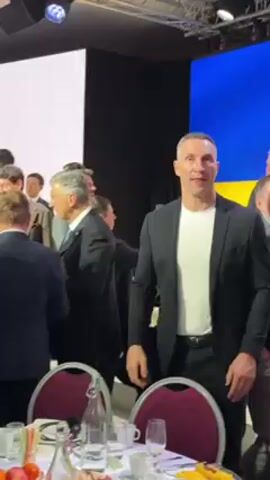 The video shows Vitali Klitschko standing amongst a crowd of people, mostly men, at an indoor event. He is wearing a black blazer over a white t-shirt, black pants, and a belt. He looks to the side and then back forward, seemingly aware of the camera. Behind him, a large screen displays blue and yellow colors reminiscent of the Ukrainian flag, partially obscured by the crowd. Several people are in suits and ties. In the foreground, there is a table set with glassware, a water bottle, and some food items, indicating a dining or reception setting. The lighting is bright, and the environment seems formal.