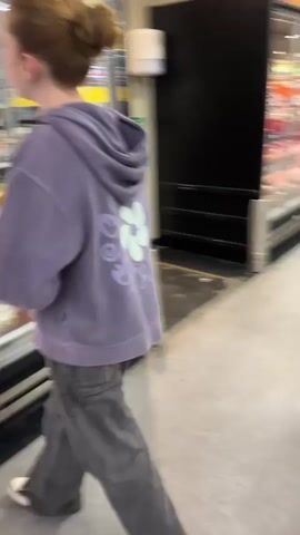 A young girl with red hair in a bun, wearing a purple hoodie and grey jeans, stands in front of a refrigerated display case in a supermarket. The case contains various food items, including what appears to be dairy products and packaged meals. To her right is another refrigerated display case filled with meat products, labeled "100% Australian Pork" and "Lamb". A young man wearing a black hoodie and blue jeans approaches the girl. He is holding a green bottle in his right hand. The girl turns and smiles at him. They appear to be having a conversation. The environment is a well-lit supermarket aisle with a concrete floor. There is a black vertical panel to the right of the meat display, and further down the aisle, another person is visible.