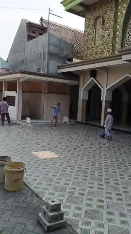 A group of young boys are playing outside a mosque. The courtyard is paved with patterned tiles. One boy in a white shirt and cap is kicking a ball towards an archway. Another boy in a light blue shirt is running and appears to be chasing the ball. A third boy in a pink shirt is standing near a covered walkway. A fourth boy is partially visible entering a doorway within the walkway. In the foreground, there are two buckets and some stacked concrete blocks. The mosque building has decorative tilework above the arches.
