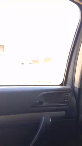 The video appears to be filmed from inside a vehicle, looking out the passenger side window. The view is of a street or outdoor area with buildings under construction. There are palm trees and other vegetation visible. In the foreground, the interior of the car is visible, including the door panel, window controls, and door handle. In the background, a person is seen carrying a box. There are also stacks of what appear to be bags of cement or sand. The sky is clear and blue, indicating daylight. The overall impression is of a somewhat arid or developing environment.