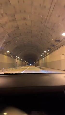 The video is filmed from the perspective of someone driving through a tunnel. The tunnel is illuminated by rows of lights along the ceiling and walls. The tunnel walls are a neutral color, and the ceiling has a textured, possibly concrete, surface. There are ventilation fans visible on the ceiling of the tunnel. The road ahead is marked with white lines. A blurred object, likely a finger or hand, is intermittently visible in the foreground of the frame. The video captures the progression through the tunnel, with the lighting and the tunnel structure being the primary focus.