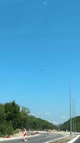 This is a series of three vertical video clips. The first clip shows a clear blue sky with an airplane visible in the distance. Below the sky, there is a road under construction with barriers and piles of dirt. Trees and foliage line the side of the road. In the second clip, the camera pans up to focus on a tall, thin pole that extends out of frame. The airplane from the first clip is now visible to the left of the pole. The third clip is identical to the first clip.