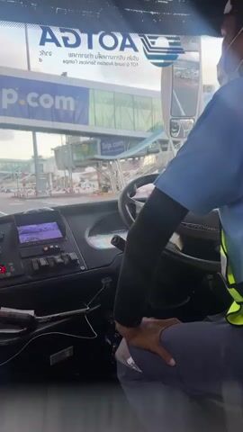 The video shows the interior of a bus from the perspective of the driver or a passenger in the front. The driver, wearing a blue uniform and a face mask, is seated behind the steering wheel, operating the vehicle. The dashboard of the bus is visible, with various buttons, screens, and indicators. Outside the bus, an airport environment is depicted. A jet bridge is visible, along with airport buildings and other vehicles. The bus appears to be moving along a road within the airport premises. The sky is cloudy, suggesting it might be dawn or dusk. The overall scene conveys a sense of movement and transportation within an airport setting.