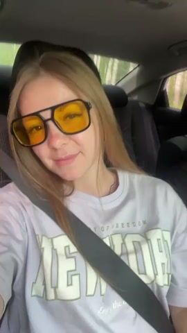 The video opens with a close-up shot of a young woman with blonde hair and yellow-tinted glasses. She is wearing a light purple t-shirt with text on it and a seatbelt. The camera then pans to reveal a man in the driver's seat, also wearing sunglasses. He is looking out the window to the right. The car appears to be in motion, with trees and foliage visible outside the windows. The video is set in the daytime, with natural light filling the car's interior. The woman smiles at the camera briefly before the video ends.