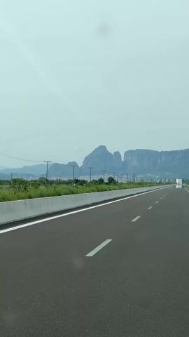 The video shows a straight, multi-lane highway under a cloudy sky. The road surface is dark and marked with white lines. A concrete barrier runs along the left side of the road. On the left, beyond the barrier, is green vegetation. In the distance, a mountainous landscape is visible, with a row of utility poles lining the horizon in front of the mountains. A truck can be seen driving far ahead on the highway in the right lane. The video is taken from the perspective of someone driving on the highway.