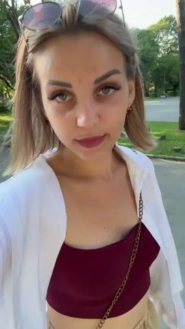 A young woman with blonde hair and makeup, wearing a white open shirt over a burgundy top and beige shorts, walks towards the camera. She is outdoors in a park-like setting with trees and grass visible in the background. The sun is shining and appears to be setting, casting a warm light. She has sunglasses perched on her head. The camera appears to be held by the woman herself, suggesting a selfie video. She looks directly at the camera with a slight smile.