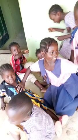 The video shows a group of seven young children, appearing to be of primary school age, gathered together indoors. They are in a brightly lit space, with a light-colored wall and a darker, possibly wooden, surface in the background. The children are dressed in various casual and school-like clothing. Several of the children are looking directly at the camera with smiles, suggesting they are posing for a picture or being filmed. One child in the foreground is wearing a striped shirt and has a wide, open-mouthed smile. Another child in the middle is wearing a pink and blue school uniform, with braided hair, and is also smiling. The overall atmosphere appears joyful and playful. The camera is positioned at a low angle, close to the children.