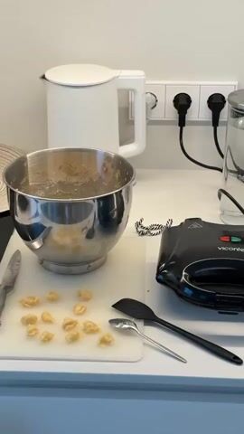 The video shows a kitchen countertop with various items for cooking. A silver mixing bowl with batter is on the left, next to a white cutting board with small, light yellow dough pieces. A black waffle maker is in the center, plugged into an outlet on the white wall. To its right, a white espresso machine stands beside a white plate filled with golden-brown, shell-shaped cookies. A glass pitcher of water is between the waffle maker and the espresso machine. A white electric kettle is in the background on the left. A black spatula and a silver spoon rest on the cutting board.
