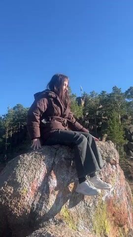 A young woman is sitting on a large, colorful rock against a clear blue sky and a backdrop of pine trees and other rocks. The woman has long, dark hair and is wearing a brown puffer jacket, dark-colored wide-leg jeans, and light-colored sneakers. She looks at the camera and smiles slightly. The rock she is sitting on has patches of different colors, possibly from graffiti. The background includes a landscape of rocks, trees, and a few other people in the distance.