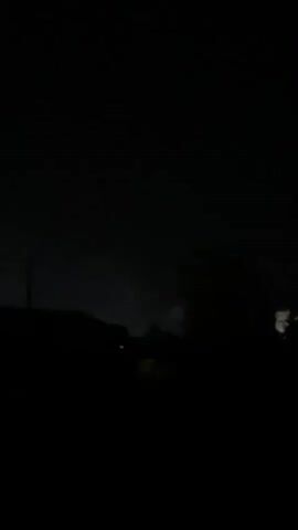 The video is a still image that appears to be taken at night. The scene is very dark, with only a few faint lights visible in the distance. The foreground is dominated by a silhouette of trees or other foliage. There is a faint glow in the sky, possibly indicating distant lightning or city lights. The overall impression is one of a rural or semi-rural setting at night, with some atmospheric activity.