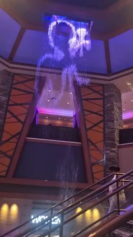 Here's a description of the video:

The video shows an indoor water feature in a large, multi-level building, likely a casino or hotel. Water flows down from a ceiling-mounted structure with blue and yellow lights, creating the illusion of a digital waterfall. The structure above the waterfall appears to have a geometric pattern.  The background includes stone pillars and a decorative wall with a brown and orange geometric design.  In the lower right corner, a person is seen riding an escalator. The overall lighting is dim with purple and pink accents highlighting upper levels of the building.