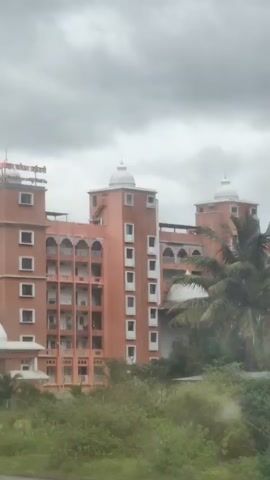 This video shows a large, ornate building with a reddish-orange facade. The building has multiple stories, domed structures, and arched windows. The sky is overcast with dark clouds. In the foreground, there is green vegetation, and the view appears to be from a moving vehicle, as indicated by slight motion blur and the way the scene pans across. Near the beginning of the video, a tree with dense foliage partially obscures the view of the building. Towards the middle, two people on a motorcycle briefly pass by. The building has signage at the top, though the text is not clearly legible. The overall impression is of a large, possibly religious or cultural institution situated in a somewhat rural or semi-urban environment under a cloudy sky.