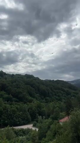 The video shows a wide shot of a densely forested mountain range under a cloudy sky. The foreground features lush green trees and foliage, with a glimpse of a red-tiled roof of a building peeking through the trees on the right. A winding road can be seen in the lower left of the frame. The middle ground is dominated by rolling hills covered in a thick canopy of trees. The background consists of more distant, hazy mountains. The sky is filled with dramatic, dark gray clouds, suggesting overcast or impending weather. There are no people or animals visible in the video. The overall impression is one of a natural, somewhat wild, and secluded landscape.