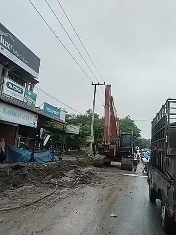 An excavator is seen on a partially wet, muddy road in front of stores. The excavator, which is a large, red-colored piece of machinery, has a filled bucket of mud raised above the ground. A small ditch is visible to the left. There's a truck to the right, partially obscuring the view. A man in dark clothing stands near the excavator. Power lines stretch across the sky above the scene.
