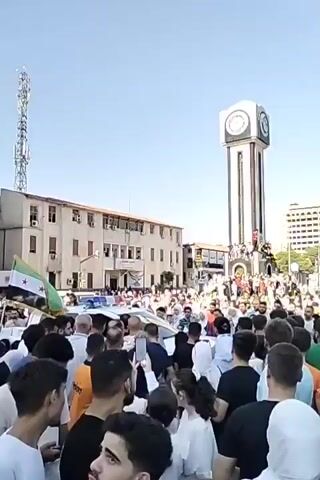 A large crowd of people has gathered in a city square under a clear, blue sky. A tall, white clock tower is prominent in the background, surrounded by the crowd. Several buildings are visible on the left and right of the square. A white police car with flashing blue lights is parked amidst the crowd. Some people are smiling and taking photos or videos with their phones. A Syrian flag is visible in the hands of someone in the crowd. The overall atmosphere suggests a public celebration or demonstration.