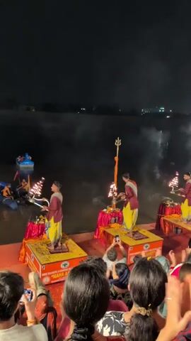 The video captures a nighttime ceremony taking place on a platform near a body of water. Several individuals dressed in traditional Indian attire, including yellow dhoti-like garments and maroon tops, are performing a ritual. They are positioned on raised, decorated platforms, each with lamps and other ceremonial items. The attendees, facing the performers, are visible in the foreground, with many holding up phones to record the event. Smoke or mist rises from the water and around the performers, illuminated by artificial lights. In the background, lights from buildings and possibly boats are reflected on the water's surface. The overall atmosphere appears to be one of reverence and cultural observance.