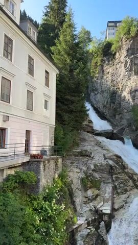 The video shows a cascading waterfall flowing down a rocky gorge, with lush green trees and vegetation on either side of the cliffs. A building, possibly a hotel or residential structure, is visible on the left side of the frame, with a cream-colored facade and multiple windows. Another building with a darker facade is perched on top of the cliff on the right. The water is white and frothy, indicating a strong flow, and the sunlight casts shadows and highlights on the rocks and trees. The scene appears to be a natural landscape enhanced by human structures.