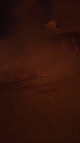 The video shows a close-up view of a barbecue grill with glowing red embers and smoke rising from it. A person's legs and feet, wearing light-colored shoes, are visible in the upper left corner of the frame, suggesting they are standing near the grill. The ground around the grill appears to be dirt or sand, with some debris scattered on it. The lighting is dim, indicating it might be nighttime or an indoor setting with low light. The camera is positioned at a low angle, looking down at the grill.