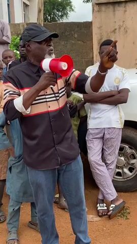The video shows an outdoor scene where a man is speaking into a megaphone. He is wearing a black cap, a collared shirt with horizontal stripes in beige, red, and white, and blue jeans. He has a beard and is gesturing with his left hand while holding the megaphone in his right. Several younger people are standing around him, listening. To the right of the man, a young man in a white t-shirt and light purple pants stands with his arms crossed. To the left and behind the speaker, other young people are visible, some wearing traditional clothing. The ground appears to be dirt. A building is visible in the background to the left, and a wall to the right. A vehicle is partially visible behind the young man on the right. The overall impression is that the speaker is addressing a group of people in a community setting.