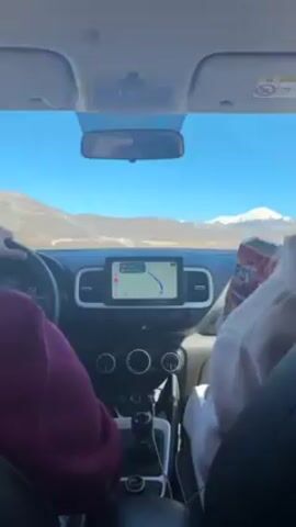 The video, shot from the front passenger seat of a car, shows two people on a road trip. The driver, wearing a maroon shirt, is focused on the road. A passenger in a white sweatshirt holds a bag of "PSY" brand chips and a cell phone. A GPS navigation display is in the car's dashboard. The video’s scenery includes mountains and a clear, blue sky.