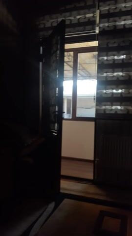 The video opens with a low-light view inside a room, looking towards a window. In the immediate foreground, a dark, ornate metal structure, possibly part of a bed frame or headboard, is partially visible at the bottom of the frame. Above this, a window with multiple panes and a wooden frame is the central focus, letting in a bright, rectangular expanse of light from outside.

To the left of the window, striped curtains with alternating dark and light vertical patterns are drawn, partially obscuring the view. To the right of the window, a dark wall or piece of furniture is visible. Through the window, an outdoor scene can be glimpsed, showing what appears to be part of a building or structure with light-colored walls and dark linear elements, possibly railings or window frames of another dwelling.

As the video progresses, the camera slowly tilts upwards, causing the ornate metal structure in the foreground to recede further down the frame. This movement brings more of the striped curtains and the upper section of the window into view, as well as a slightly different perspective of the bright outdoor area. The room itself remains dim throughout the footage, creating a strong contrast with the brightly lit exterior visible through the window. The camera then slowly tilts back down towards its initial position, again highlighting the ornate foreground object and the lower portion of the window. This gentle up-and-down panning motion continues for the duration of the video, maintaining the focus on the window and its immediate surroundings. No people or visible text are present in the video.