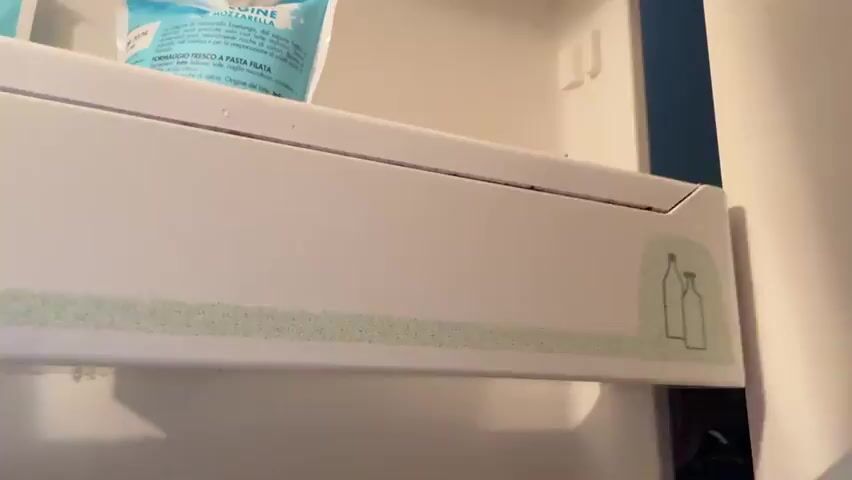 The video opens with a close-up view inside a refrigerator, showing a white shelf with some blue packaging, possibly food items, and a decorative band with bottle outlines. The scene then transitions to a dark, almost black frame, possibly indicating a moment of darkness or a transition.

The camera then returns to the refrigerator interior, showing the same shelf with the blue packaging. Suddenly, a man appears in the frame, leaning into the refrigerator. He is shirtless, revealing tattoos on his arm and chest, and has short, styled brown hair. He smiles directly at the camera and waves.

The background suggests the man is in a room that appears to be a kitchen or living area, with furniture and windows visible. The overall mood seems casual and friendly, with the man greeting the viewer. The video appears to be a short, informal recording, likely a personal message or greeting.

There is some visible text on the blue packaging in the refrigerator, but it is partially obscured and difficult to read clearly. The setting appears to be indoors, with artificial lighting. The time of day is not discernible. The location context provided is Sesto Calende, Italy.