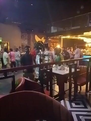 The video depicts a film or television shoot taking place in a nightclub or bar setting. A camera crew is positioned amidst a crowd of people, presumably actors and extras. The camera is mounted on a tripod and is surrounded by crew members, some of whom are operating equipment. The background features bar tables with glasses, a bar with colored lighting, and a large golden bird sculpture on the wall. Signage for "Kingfisher Ultra" is also visible. The lighting is low, typical of a nightclub, with some spot lighting for the shoot. People are milling around and appear to be either waiting for direction or acting.