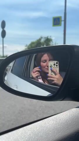 The video shows a young woman with long brown hair and bright blue fingernails taking a selfie in a car's side mirror. She is wearing a light-colored top. She is holding a pink scrunchie in her left hand and a smartphone in her right hand. The phone is in a beige case with a black camera module. The woman is smiling and looking at the phone. The side mirror reflects her face and upper body. Outside the car, there is a blue sky with some clouds, trees, and a traffic sign with a yellow diamond shape. The car appears to be moving, as indicated by the slight blur in the background. The overall scene is casual and everyday.