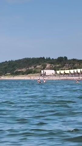 The video shows a beach scene from the water's perspective. The water is a choppy blue. In the distance, a sandy beach is lined with small white cabins with yellow awnings. People are visible on the beach and in the water, some swimming and others relaxing. The beach is backed by a sloping, tree-covered hill. The sky is clear and blue.