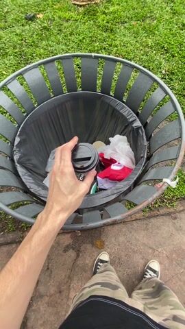 The video starts with a first-person view of a person dropping a used coffee cup into a trash can. The person then walks along a sidewalk, transitioning into a wider view of a city square with pigeons. A person is sitting on a bench surrounded by pigeons, with urban traffic visible in the background.