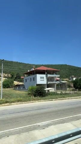 This is a vertical, outdoor video shot from a moving vehicle. The video shows a roadside view of a village nestled on a hillside under a clear blue sky. A paved road runs horizontally across the bottom of the frame. On the left side of the road, there is a multi-story building with a red-tiled roof, white exterior walls, and a balcony. To the right of this building, there are several smaller houses with red-tiled roofs, interspersed with trees and greenery. A telephone pole with wires is visible on the right side of the road, next to the houses. The hillside behind the village is covered in green vegetation. The lighting suggests it is daytime, likely late morning or early afternoon.