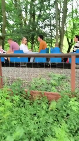 A miniature train with an orange, yellow, and blue engine and multiple blue passenger cars moves from left to right on a gravel track through a lush, green, wooded area. Several adults and children are seated in the open-top blue cars, looking forward and to the sides. The train emits a horn sound as it passes. A metal fence with brown wooden posts and green leafy vegetation runs along the foreground, separating the viewer from the train track. Large trees with green leaves frame the scene.