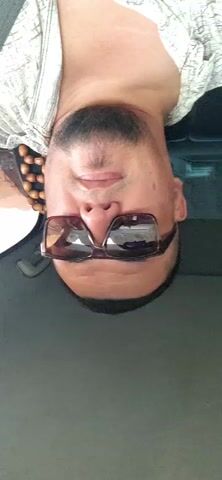 A man wearing sunglasses and a patterned shirt is seen upside down in the video. He has a goatee and is looking directly at the camera. The background appears to be the interior of a car, with a seat and a window visible. The lighting suggests it is daytime.