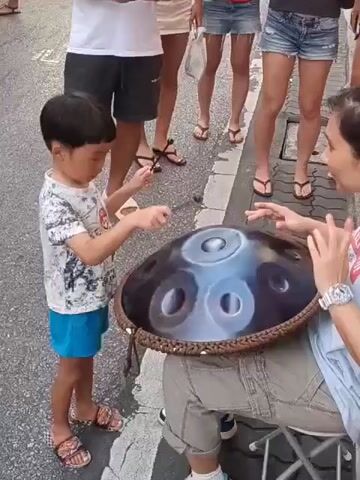 The media file captures a street scene in Phuket City Municipality, Thailand, likely during the day given the bright lighting. The foreground features a young boy, dressed in a patterned t-shirt and blue shorts, interacting with a woman playing a handpan drum. The boy holds a stick, seemingly ready to play or mimicking the woman's actions. The handpan is a large, dark, metallic instrument with circular indentations. The woman, wearing a blue shirt, is seated and appears to be demonstrating or performing with the instrument.

Behind them, a group of people stand, some observing the performance. They are casually dressed, with some wearing shorts and t-shirts, suggesting a warm climate. The setting appears to be a public street, possibly a pedestrian area with shops or stalls lining the sides, hinted at by the white umbrellas and decorative floral arrangements visible further down the street. One man in the background wears a white t-shirt with the text "BANGKOK PANTHERS" and what appears to be a logo. The overall mood seems relaxed and engaging, with a sense of shared experience between the performers and the onlookers. The people's expressions and body language suggest interest and enjoyment of the music.