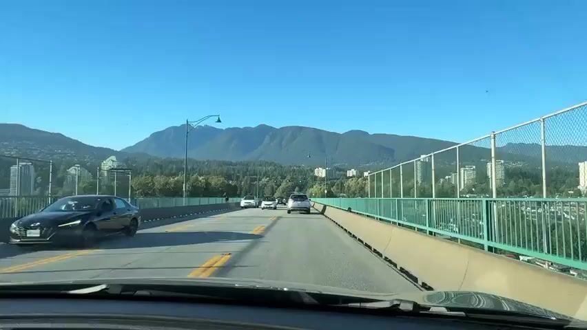 A first-person view from a vehicle traveling on a bridge on a clear, sunny day. The road ahead has two lanes in each direction, separated by double yellow lines. Other vehicles, including cars and SUVs, are visible on the road. The bridge has a green railing on the right side. Beyond the railing, a cityscape with tall buildings and trees can be seen. In the distance, a range of mountains with a clear blue sky overhead forms the backdrop. Traffic lights are visible on the bridge, displaying red and green signals. The vehicle appears to be moving at a moderate speed.