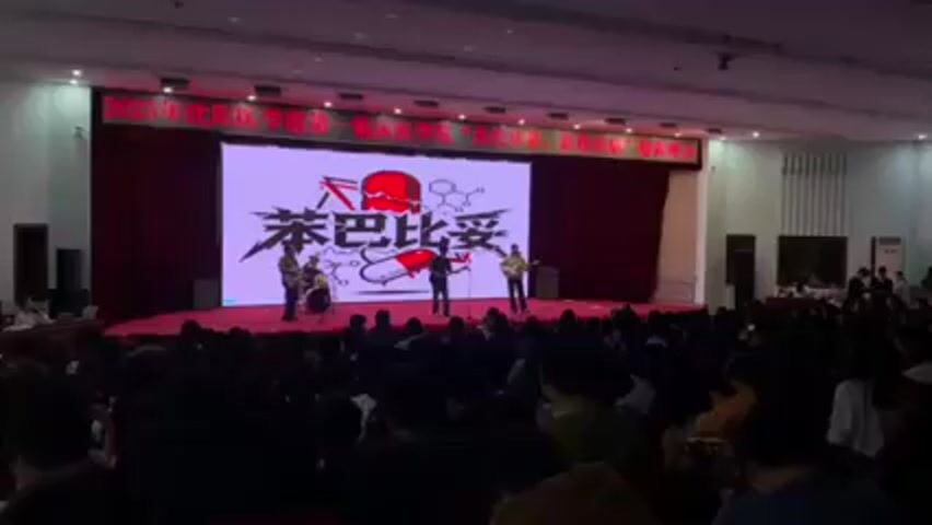 A band performs on stage for a large audience in an auditorium. The stage has a large screen displaying a graphic design with Chinese characters. The band consists of a drummer, a bassist, and two guitarists, one of whom is singing into a microphone. The audience is seated and facing the stage. The auditorium is well-lit.