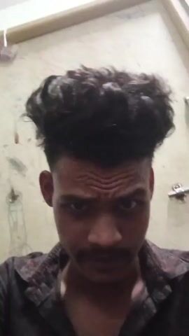 The video shows a close-up of a young man with dark, curly hair and a mustache. He is wearing a dark patterned shirt. In the first few seconds, he is seen touching his hair. The background appears to be a wall with some writing on it, and a light fixture. The lighting is somewhat dim.