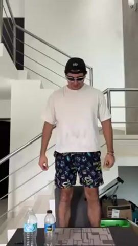 A young man is standing in front of a table in an indoor space. He is wearing a white t-shirt, patterned shorts, a black baseball cap, and sunglasses. He is tapping his thigh rhythmically. Behind him is a modern staircase with metal railings and a white wall. On the table in front of him, there are two water bottles and what appears to be a geometric design. In the background, there is a cardboard box and some exercise equipment. The room is brightly lit.