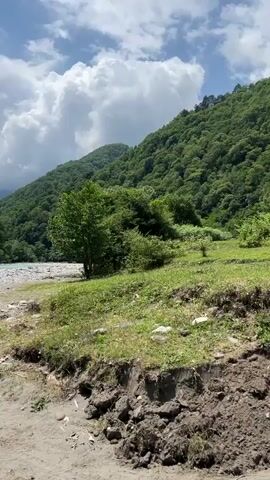 This media file is a video showcasing a scenic natural landscape. The primary subject is a wide, turquoise river flowing through a lush, green valley. The riverbanks are lined with smooth, grey pebbles and some sandy areas. On the right side of the frame, a steep, tree-covered mountainside rises dramatically, its dense foliage a vibrant green. In the distance, further mountains are visible, some appearing hazy in the background. The sky above is a bright blue, dotted with fluffy white clouds, suggesting a pleasant, possibly warm day.

The video appears to be a passive observation of the environment, likely a walk or a stationary view from the riverbank. There are no people or specific objects visible, emphasizing the untouched, natural beauty of the setting. The time of day seems to be daylight, given the brightness and shadows. The weather appears fair, with sunshine indicated by the bright sky and well-lit landscape. The location, identified as Bzypta, Abkhazia, implies a mountainous, riverine region. The overall mood conveyed is one of tranquility and natural splendor. No text is visible in the media file.