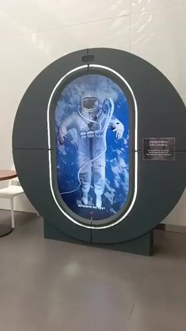 A video showcases a display of an astronaut in a spacesuit floating against a backdrop of Earth from space. The astronaut is wearing a white spacesuit with the letters "СССР" visible on the helmet. A sign on the display reads "УВАЖАЕМЫЕ ПАССАЖИРЫ" which translates to "Dear passengers". Below the astronaut's image, text in Russian reads "Встаньте на круг," meaning "Stand on the circle." A red arrow points downwards towards a circular area marked on the floor. The display is oval-shaped and illuminated by white LED lights around its perimeter. The entire setup is set against a plain, light-colored wall.