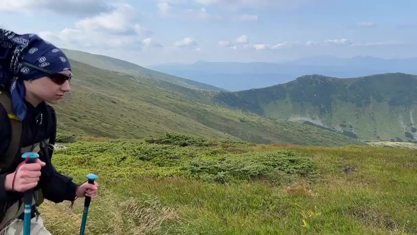 A group of hikers walks along a trail in a mountainous landscape. The first hiker is a person wearing a dark jacket and a bandana, carrying a large backpack and using trekking poles. The second hiker is visible from the chest up, wearing a hooded jacket and a baseball cap, also with a large backpack and trekking poles. The third hiker is a person in a green jacket and black pants, using trekking poles and wearing a backpack. The landscape features rolling green hills with patches of grass and low-lying shrubs, and in the background, there are more distant mountains under a cloudy sky.