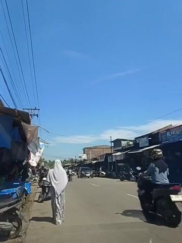 A person wearing a white headscarf and a patterned dress walks down a paved road. To the left of the road are market stalls with blue tarps covering them. Several motorcycles are parked on the left side of the road. On the right side of the road are buildings with dark roofs and windows. The sky is clear and blue with some wispy clouds. There are power lines running across the top of the frame. Other vehicles, including motorcycles and cars, are visible on the road in the distance. The overall scene depicts a busy street market on a sunny day.