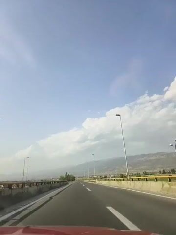 The video shows a first-person view of a car driving on a highway. The sky is blue with scattered clouds. On either side of the road are barriers and streetlights. In the distance, there are mountains and some vegetation. The road appears to be in good condition with clear lane markings. The sun is shining, casting shadows. There are no other vehicles or people visible in the immediate vicinity of the car. The overall environment suggests a daytime drive in a semi-rural or suburban area.